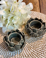 Two small dark grey artichoke shaped tea lights, pictured on a white rattan placemat.