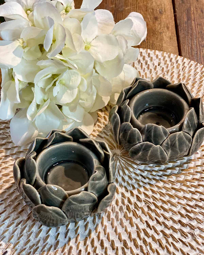 Two small dark grey artichoke shaped tea lights, pictured on a white rattan placemat.