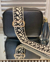 Black tassel bag with a beige/black print bag strap going across it. On a mirrored table.