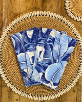 Set of four napkins printed with blue banana leaf print, on rattan shell placemat.