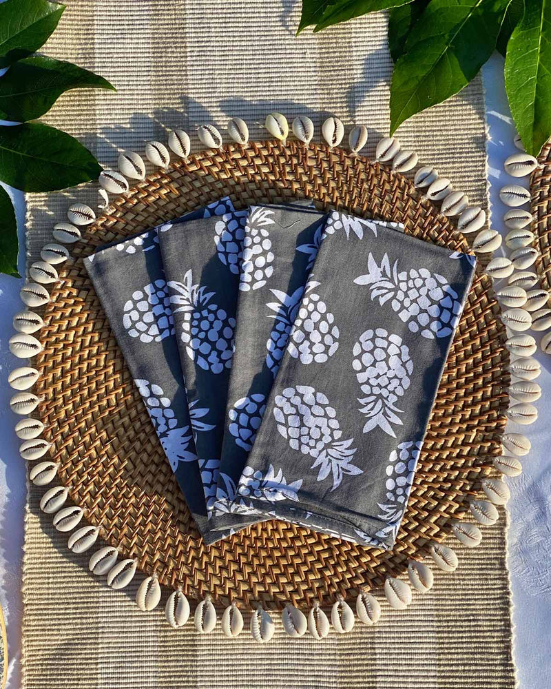 Set of four grey napkins printed with white pineapples, on rattan shell placemat.
