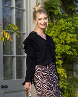 A long sleeved black blouse adorned with subtle frills on the shoulders and sleeves. She is smiling outside by a floor length bay window.