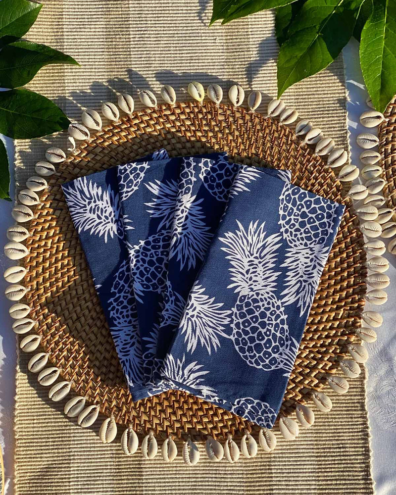 Set of four navy napkins printed with white pineapples, on rattan shell placemat.
