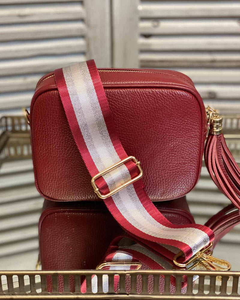 Red tassel bag with red/silver stripe bag strap going across it. On a mirrored table.