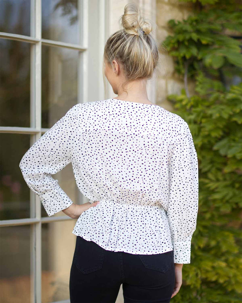 Back pose of a woman by a terrace door wearing a black dotted print on white. With a elasticated waist and cross over neck line.