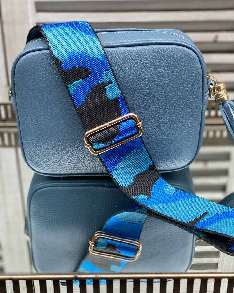 Denim blue tassel bag with a black/two blue camo print bag strap going across it. On a mirrored table.