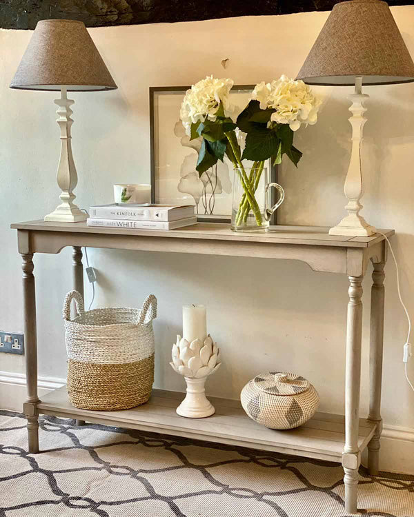 A wooden grey console table with one shelf at the top and one shelf at the bottom. It has two lamps either side and homeware accessories.