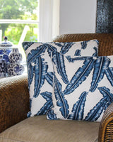 Two cushion covers printed with navy palm leaves on white cotton. Pictured on a rattan chair.