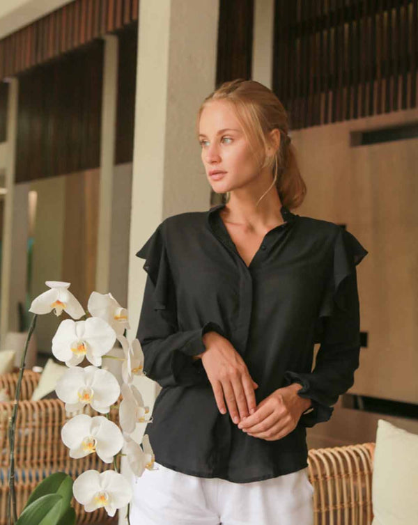 Model is wearing a long sleeved black blouse adorned with subtle frills on the shoulders and sleeves. She is inside a house in Bali.