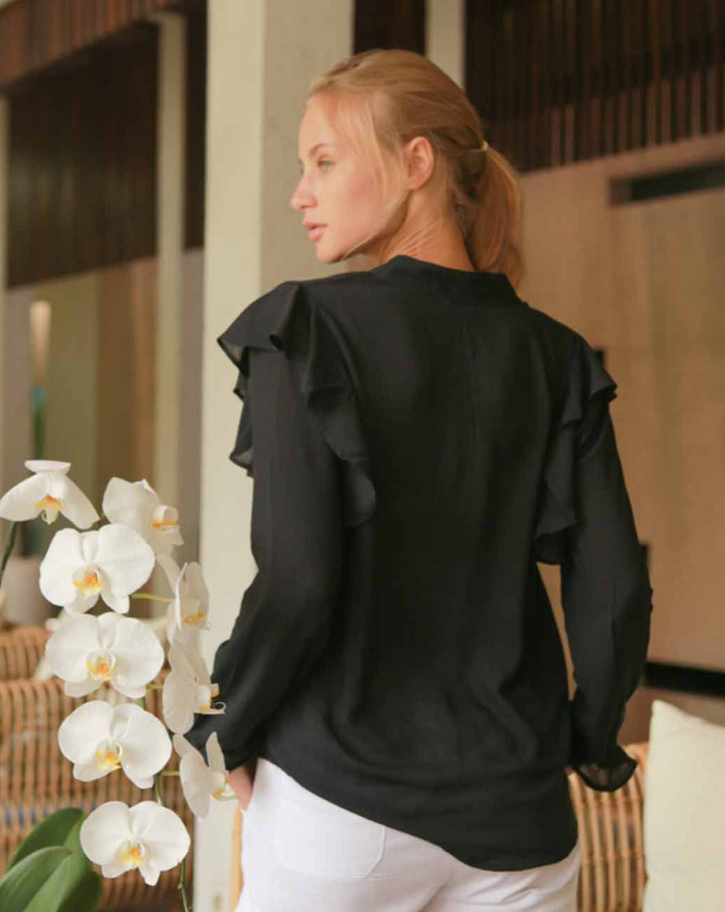 Behind shot of model who is wearing a long sleeved black blouse adorned with subtle frills on the shoulders and sleeves. She is inside a house in Bali.